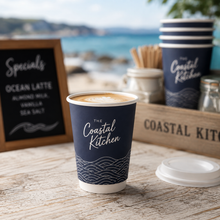 Load image into Gallery viewer, A navy blue custom branded hot cup with white script reading 'The Coastal Kitchen' and a wave pattern near the bottom, filled with coffee and placed on a wooden table. A matching cup stack, lid, and a beach-themed chalkboard sign are visible in the blurred background.
